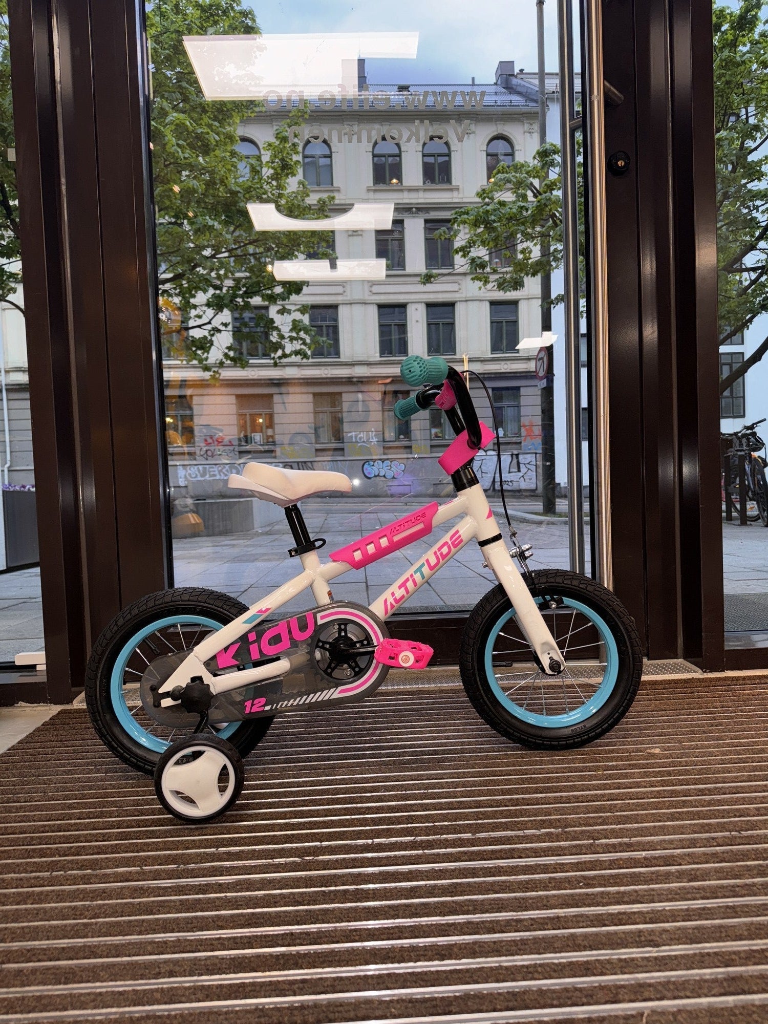 Hvit og rosa Mechanic Arts Snurr barnesykkel i butikkvindu, sidevisning med gatemotiv – lett balanse sykkel for små barn