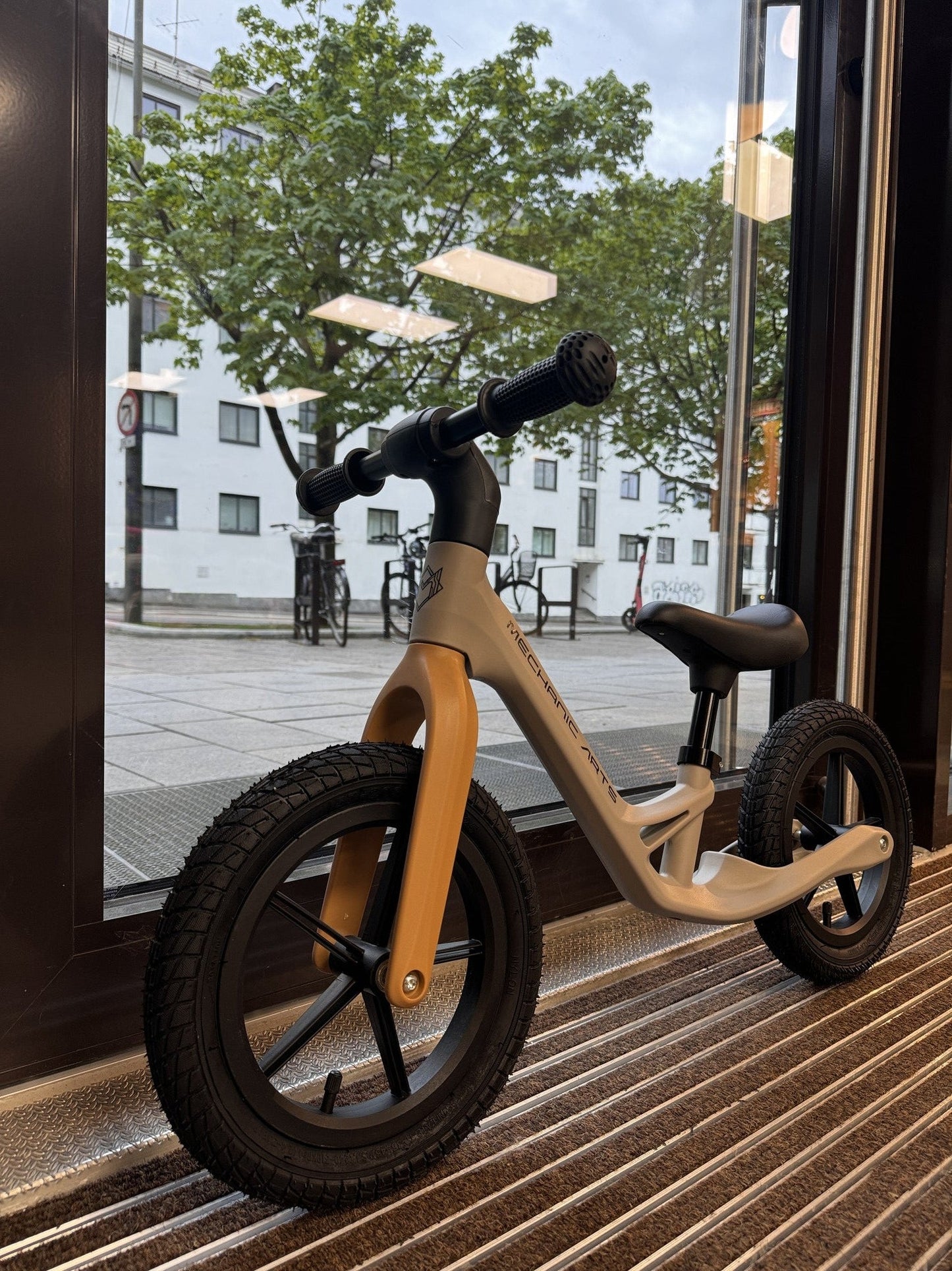 Mechanic Arts Trillr beige barnesykkel med vinklet sidevisning i butikkvindu – lett balanse sykkel for små syklister