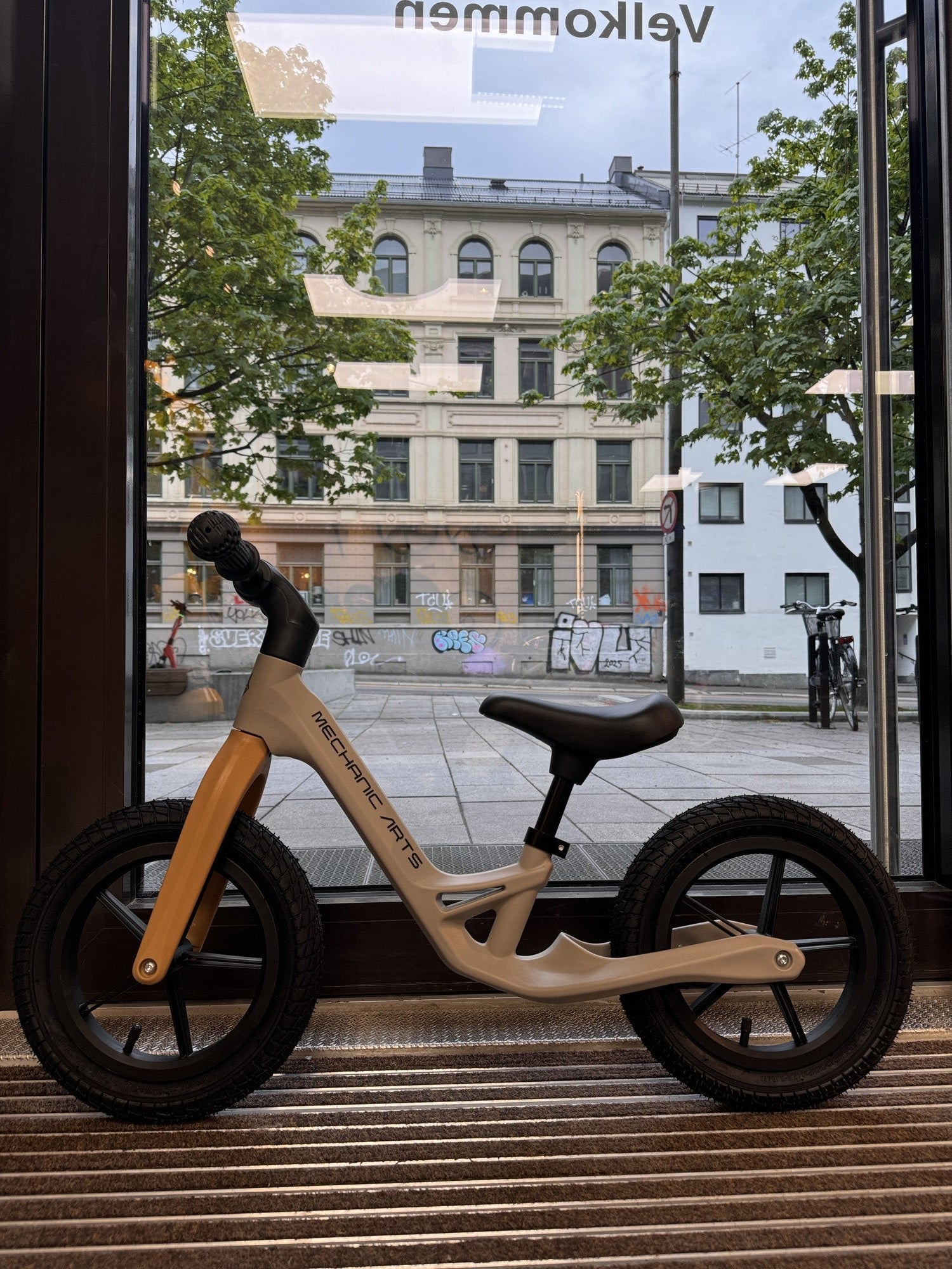 Beige balanse sykkel for barn fra Mechanic Arts Trillr, vist i butikkvindu fra siden – en stilig barnesykkel uten pedaler